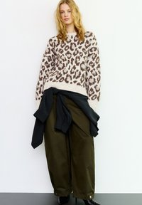 Bézs és barna leopárdmintás kötött pulóver, laza olívazöld nadrággal párosítva. Fekete inget a deréknál megkötve; fényes fekete cipő.