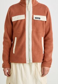 Pomarańczowa polarowa kurtka z kremowymi akcentami, z pełnym zamkiem błyskawicznym, wysokim kołnierzem, dwiema przednimi kieszeniami i logo marki na lewej części klatki piersiowej.