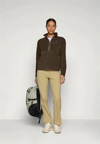 Kvinne som har på seg en brun fleecegenser, beige bukser, hvite joggesko, og holder en grå og svart ryggsekk mot en ensfarget lys bakgrunn.