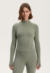 Priliehavá bunda s dlhými rukávmi v tlmenej zelenej farbe. Vyrobená z elastického materiálu s vysokým golierom a zipsovým zapínaním. S otvoromi na palce na koncoch rukávov.