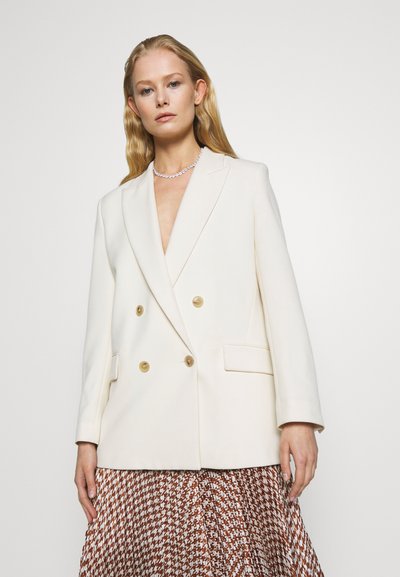 Blazer blanc à double boutonnage en tissu lisse avec des boutons dorés et des revers crantés, associé à une jupe brune à motifs.