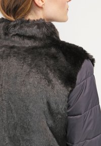 Damska odzież wierzchnia: ciemny kołnierz z futra syntetycznego o miękkiej fakturze, zestawiony z szarym rękawem puchowym. Gładki materiał, funkcjonalny design.