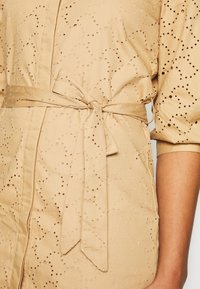 Vestido wrap beige con un patrón texturizado y agujeros recortados, un cinturón atado en la cintura y mangas cortas con fruncidos.