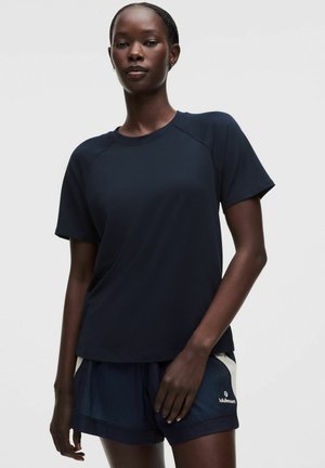 Vrouw met een donkere huid die een marineblauw shirt met korte mouwen en marineblauwe sportshorts draagt, staand tegen een effen lichte achtergrond.
