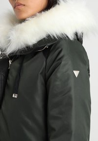 Giacca a vento verde con cappuccio in pelliccia ecologica bianca, dotata di cerniera e tasca laterale. Include un dettaglio logo metallico sulla manica.