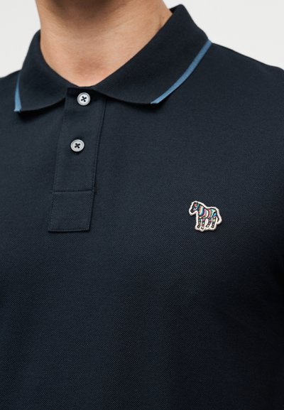 Μπλε ναυτικό πόλο με ριμπ ύφασμα στον γιακά με ανοιχτό μπλε λεπτομέρειες, δύο κουμπιά και ένα πολύχρωμο κεντητό λογότυπο ζέβρας στην αριστερή πλευρά.