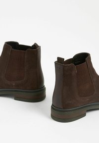 Las botas de tobillo de ante marrón cuentan con paneles laterales elásticos y acentos texturizados. El diseño incluye una suela resistente y detalles de costura.