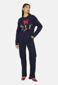 Maglione blu navy con lettere colorate in paillettes che formano la parola "ELF", con fiocco rosso e dettagli a stella, abbinato a jeans scuri e scarpe rosse.
