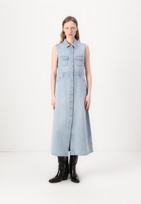 Lyseblå denim-sansermiddress med knappp foran, to brystlommer og en litt utsvingt skjørt; brukt med svarte knelang støvler.