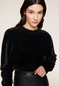 Svart mjuk tröja med rund halsringning och långa ärmar, kombinerad med en svart läderkjol med ett bälte i guldfärgade detaljer.