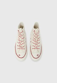Αθλητικά παπούτσια με ψηλό πάνω μέρος από κρεμ καμβά με κόκκινα σχέδια καρδιών στα κορδόνια, κόκκινες λαστιχένιες λεπτομέρειες και λογότυπο καρδιάς στη μύτη.