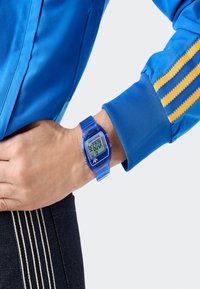 Person i blå jacka med gula ränder och digital blå armbandsur står med handen på höften.