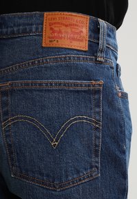 Primer plano de unos jeans Levi's 501 azules que muestra el bolsillo trasero con el característico bordado en forma de arco y la etiqueta de cuero de la marca encima de la cintura.