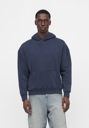 Homme face à l'avant portant un sweat à capuche bleu foncé et un jean bleu clair avec des motifs d'étoiles sur la partie supérieure de la cuisse sur un fond uni.