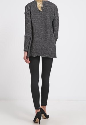 Kvinde stående med ryggen til, iført en tekstureret mørkegrå langærmet top, sorte leggings og sorte højhælede sko.