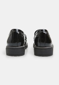 Zapatos de charol negro con punta redonda y una suela negra texturizada y baja, presentando una correa y un hebilla en la parte superior.