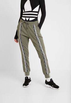 Person trägt olivgrüne Adidas-Jogginghosen mit schwarzen und weißen Logo-Streifen, schwarze Adidas-Sneaker und ein schwarzes Langarmshirt mit großem weißem Logo.