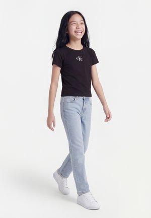 Νεαρή κοπέλα που περπατά χαμογελαστή, φοράει μαύρο μπλουζάκι Calvin Klein, ανοιχτόχρωμα μπλε τζιν και λευκά αθλητικά παπούτσια, με φόντο λευκό.