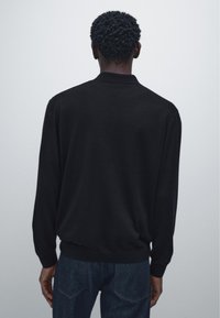 Man with short curly hair wearing a black long-sleeve shirt and dark blue jeans, standing facing away against a plain background.