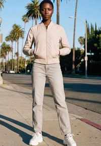 Fiatal személy bézs színű bomberdzsekiben, világosszürke farmerben és fehér tornacipőben napos utcán áll, pálmafákkal.