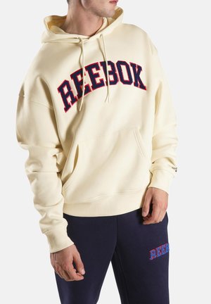 Hombre vistiendo una sudadera con capucha de color crema de Reebok con un gran logo en rojo y azul, combinado con pantalones azul marino con un pequeño logo de Reebok.