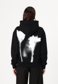 Felpa nera in cotone con un design texturizzato, caratterizzata da grafiche floreali bianche sul retro. Il cappuccio e i polsini a coste completano il design.