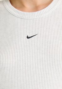 Ribbete hvit strikket skjorte med rund hals og en liten svart Nike-logo sentrert på brystet. Myk tekstur og uformell design.