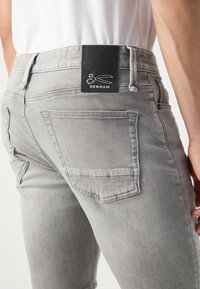 Gray denim jeans with a fitted design. Back features two pockets and a black leather label with white scissor graphic and "DENHAM" text.