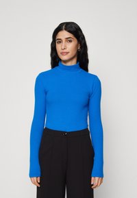 Pulover albastru cu guler înalt și mâneci lungi, dintr-un material neted și elastic, asortat cu pantaloni negri cu talie înaltă, având o siluetă ajustată.