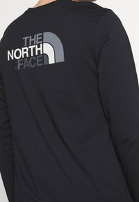 Czarna koszula z długim rękawem wykonana z miękkiego materiału, z dużym szaro-białym logo "The North Face" na plecach.