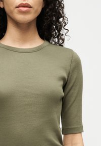 Kvinne med krøllete hår iført en olivengrønn ribbestrikket genser med korte ermer, vist fra nakken til midt på overkroppen mot en ensfarget bakgrunn.