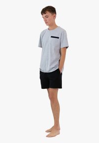 Szara bawełniana koszulka z krótkim rękawem i czarną kieszenią, w połączeniu z czarnymi szortami. Model stoi boso, z rękami w kieszeniach.