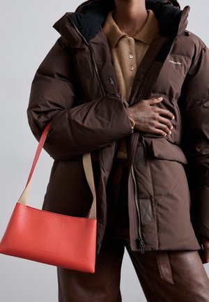 Persoon in een donkerbruine pufferjack en een beige gebreid shirt, houdt een rode leren schoudertas met beige banden vast tegen een grijze achtergrond.