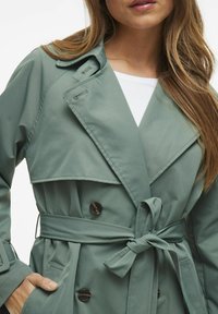 Trench coat verde chiaro con design doppiopetto, ampie patte, dettagli con bottoni e cintura legata in vita, caratterizzato da una texture liscia.