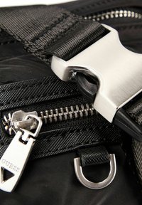 Sac en nylon noir avec une boucle métallique, un curseur de fermeture éclair portant le logo "GUESS" et une sangle texturée avec un détail en boucle.