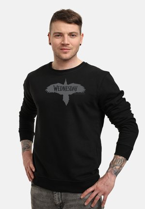 Man met kort haar en tatoeages, gekleed in een zwarte sweatshirt met "Woensdag" en een silhouet van een vogel, staand met zijn handen op zijn heupen.