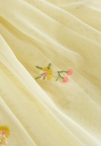 Gult gennemsigtigt stof med delikat broderede pink og gule blomster med grønne stilke på bløde folder.