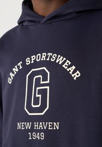 Sweat à capuche bleu marine avec texte blanc "GANT SPORTSWEAR" courbé au-dessus d'un grand "G" et "NEW HAVEN 1949" en dessous sur la poitrine.