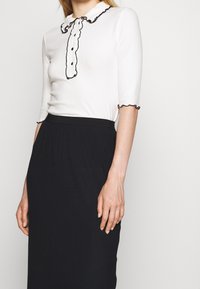 Bílý žebrovaný top s černým zvlněným límečkem a knoflíky, spárovaný s černou žebrovanou sukní. Materiál je hladký a těsně přiléhavý.