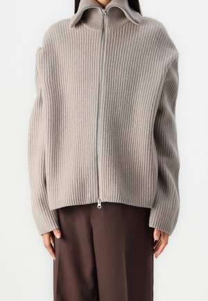 Personne portant un pull beige en maille côtelée zippé avec un col montant et un pantalon marron à jambes larges sur un fond uni.