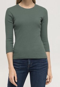 Grön ribbad långärmad topp med rund halsringning, figurnära silhuett och ärmar i medellängd. Kombinerad med mörkblå jeans.