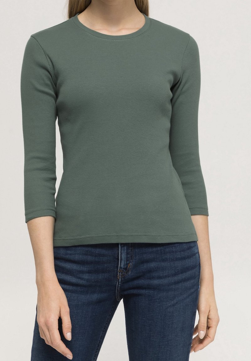 Grön ribbad långärmad topp med rund halsringning, figurnära silhuett och ärmar i medellängd. Kombinerad med mörkblå jeans.