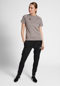 Frau steht mit den Händen in den Taschen, trägt ein graues kurzärmeliges Shirt, schwarze Sporthosen und schwarze Turnschuhe vor weißem Hintergrund.