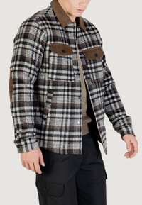 Giacca in flanella a quadri neri, bianchi e marroni, con un colletto in corduroy marrone, due tasche sul petto e una vestibilità rilassata. Tessuto texture con motivi visibili.