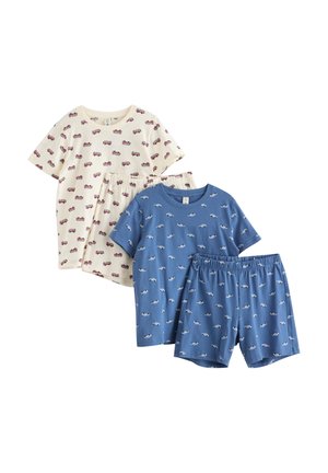 Jeune garçon debout pieds nus, portant une chemise bleue à manches courtes et un short avec de petits motifs de voitures blanches sur un fond blanc uni.