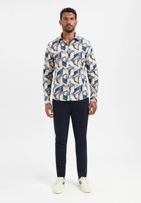 Man staat in een geometrisch patroon met lange mouwen, donkere slim-fit broek en witte sneakers met marineblauwe accenten.