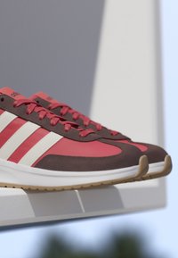Paar rote und braune Sneakers mit weißen Streifen und Gummisohlen, die auf einer weißen Kante vor einem unscharfen Hintergrund liegen.