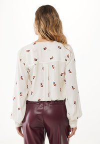 Vrouw met lang golvend haar draagt een witte blouse met kleine bloemmotieven en bordeauxleren broek, staand met de rug naar de camera.