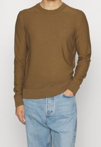 Pull marron à col rond en maille douce avec poignets et ourlet côtelés ; présente un détail logo sur la poitrine. Associé à un jean bleu clair.