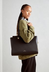 Sort struktureret tote taske med en glat tekstur, prydet med et gyldent logo. Modellen bærer en olivenfarvet quiltet jakke og viser tasken.